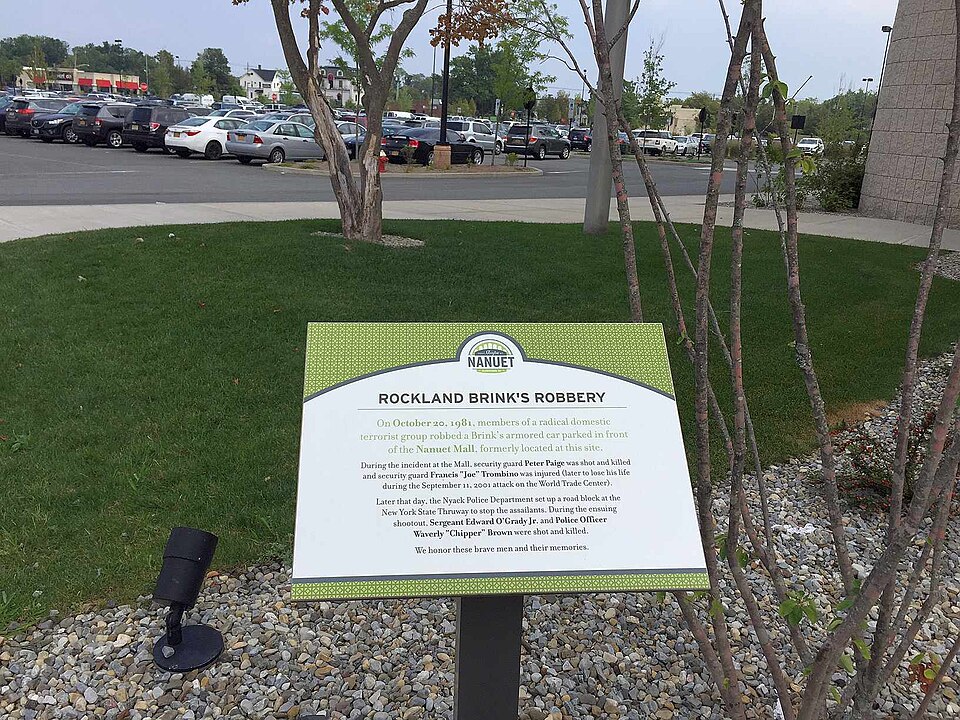 Historical marker in front of a parking lot with trees and buildings in background.